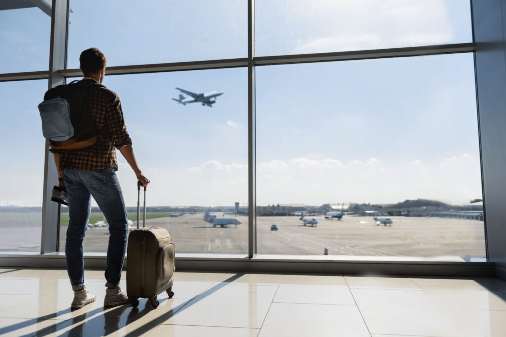 Young man is standing near window at the airport and watching plane before departure He is standing and carrying luggage Focus on his back